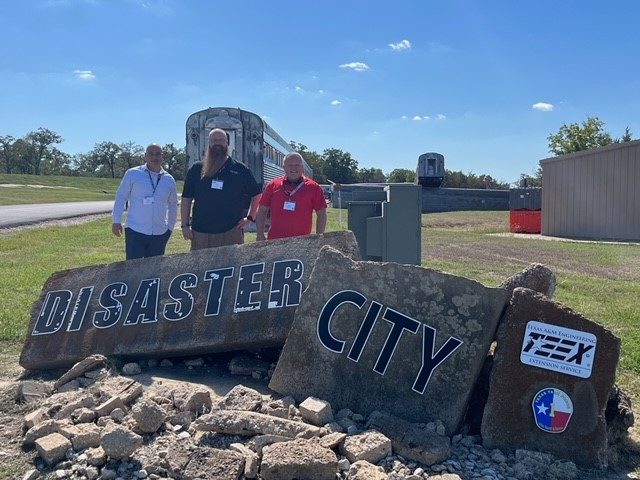 Three men stand behind large broken rocks with the words "Disaster City" painted on the rocks.